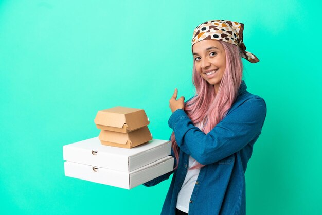 Young mixed race woman holding pizzas and burgers isolated on\
green background pointing back