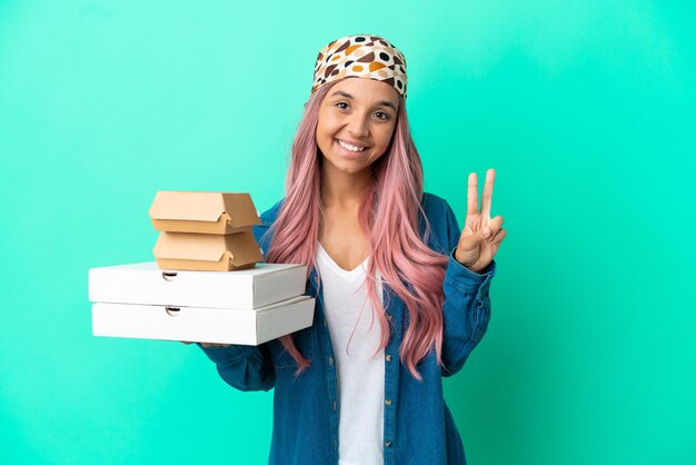 Young mixed race woman holding pizzas and burgers isolated on green background smiling and showing victory sign