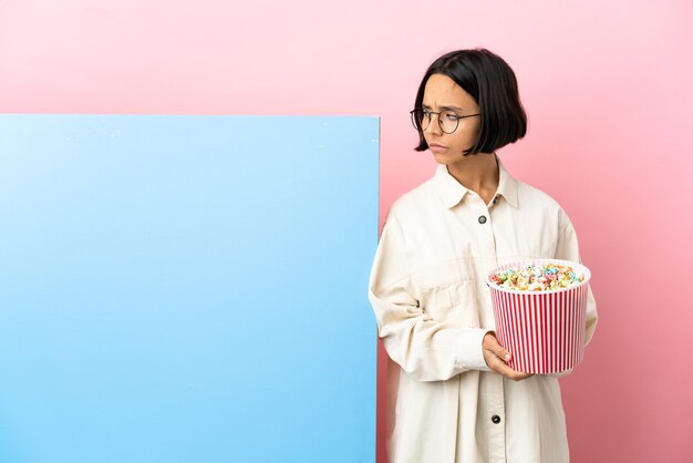 Young mixed race woman holding popcorns with a big banner over isolated background having doubts while looking side