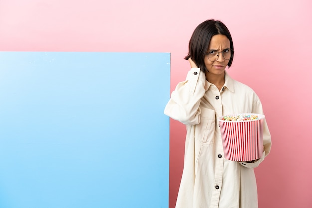 Young mixed race woman holding popcorns with a big banner over isolated background having doubts