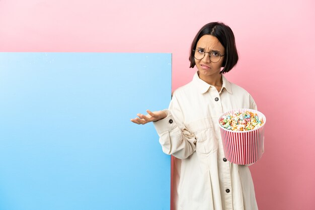 Young mixed race woman holding popcorns with a big banner over isolated background having doubts