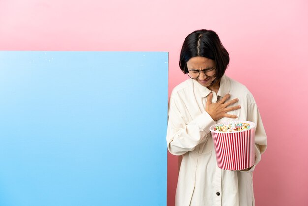 Young mixed race woman holding popcorns with a big banner over isolated background having a pain in the heart