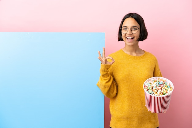 Young mixed race woman holding popcorns with a big banner over isolated background showing ok sign with two hands