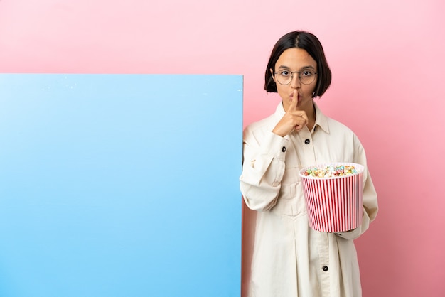 Giovane donna di razza mista che tiene i popcorn con un grande striscione su uno sfondo isolato che mostra un segno di gesto di silenzio che mette il dito in bocca