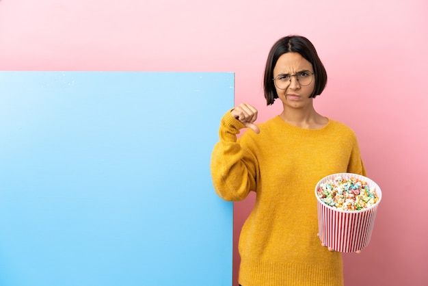 Young mixed race woman holding popcorns with a big banner over isolated background showing thumb down with negative expression