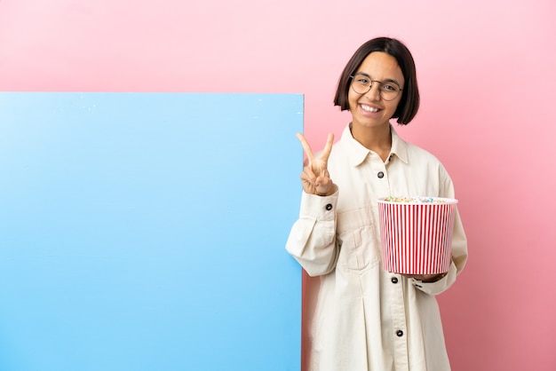 Young mixed race woman holding popcorns with a big banner over isolated background smiling and showing victory sign