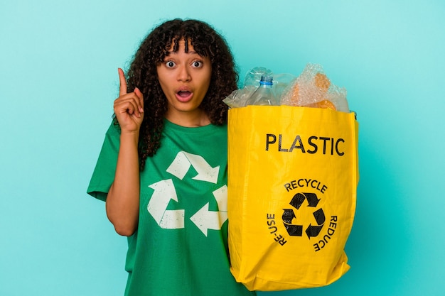 Young mixed race woman holding a recycled plastic bag isolated on blue background having an idea, inspiration concept.