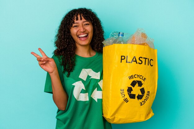 Young mixed race woman holding a recycled plastic bag isolated on blue background joyful and carefree showing a peace symbol with fingers.