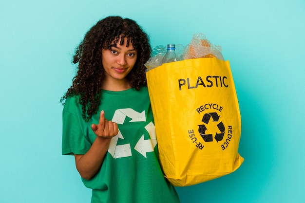 Young mixed race woman holding a recycled plastic bag isolated on blue background pointing with finger at you as if inviting come closer.