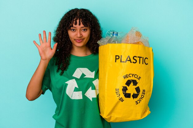 Young mixed race woman holding a recycled plastic bag isolated on blue background smiling cheerful showing number five with fingers.