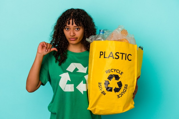 Young mixed race woman holding a recycled plastic bag isolated on blue wall showing a dislike gesture, thumbs down. Disagreement concept.