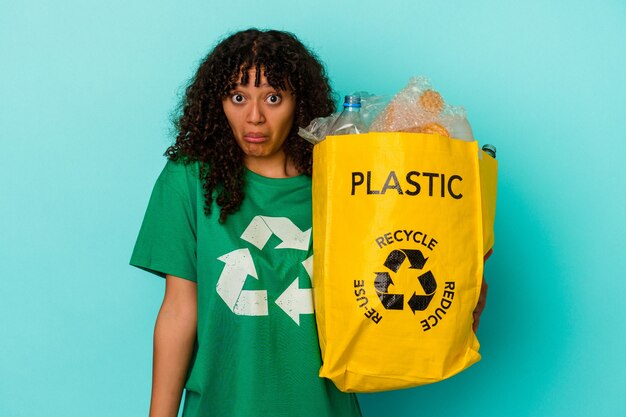 Young mixed race woman holding a recycled plastic bag isolated on blue wall shrugs shoulders and open eyes confused.