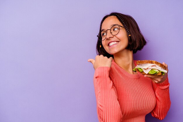 Young mixed race woman holding a sandwich isolated on purple background points with thumb finger away, laughing and carefree.