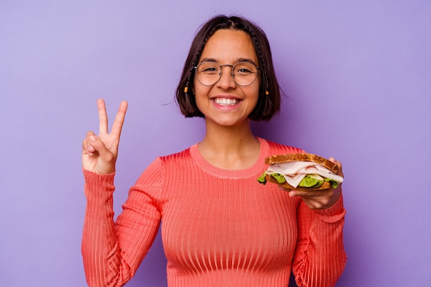 Молодая женщина смешанной расы, держащая бутерброд, изолированные на фиолетовом фоне, показывая номер два пальцами.