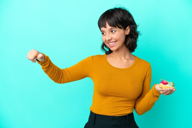 Young mixed race woman holding a tartlet isolated on blue background giving a thumbs up gesture