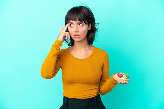 Young mixed race woman holding a tartlet isolated on blue background having doubts and thinking