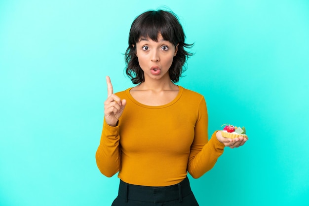 Young mixed race woman holding a tartlet isolated on blue background intending to realizes the solution while lifting a finger up