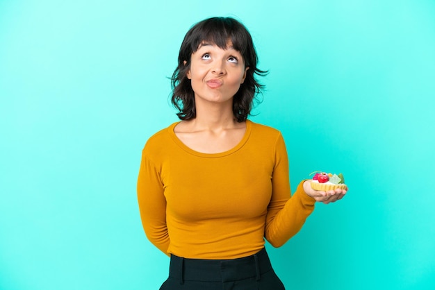 Young mixed race woman holding a tartlet isolated on blue background and looking up