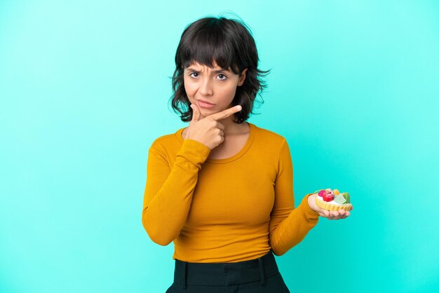 Young mixed race woman holding a tartlet isolated on blue background thinking