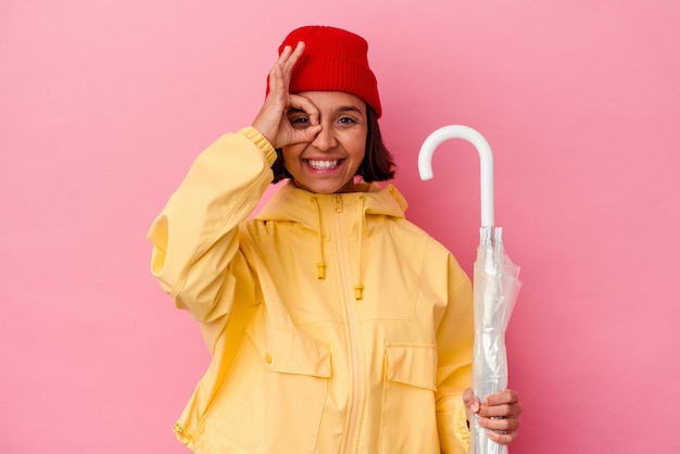 Young mixed race woman holding an umbrella isolated on pink background excited keeping ok gesture on eye.