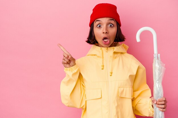 Young mixed race woman holding an umbrella isolated on pink wall pointing to the side