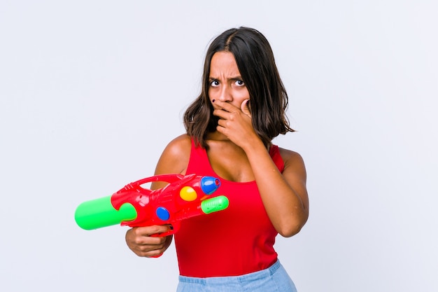 Young mixed race woman holding a water gun isolated covering mouth with hands looking worried.