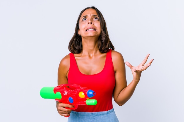 Young mixed race woman holding a water gun isolated screaming to the sky, looking up, frustrated.