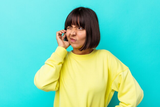 Young mixed race woman isolated on blue background covering ears with fingers, stressed and desperate by a loudly ambient.