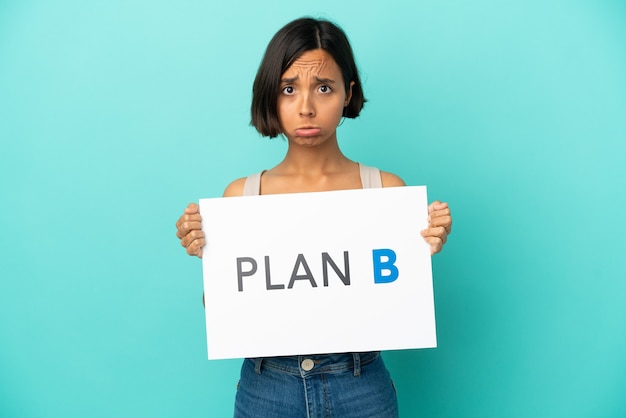 Young mixed race woman isolated on blue background holding a placard with the message PLAN B with sad expression