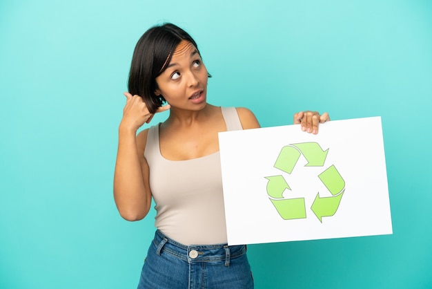 Young mixed race woman isolated on blue background holding a placard with recycle icon and doing phone gesture