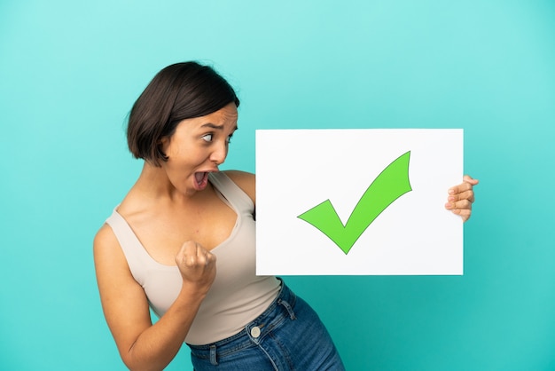 Young mixed race woman isolated on blue background holding a placard with text Green check mark icon and celebration a victory