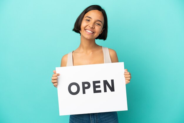 Young mixed race woman isolated on blue background holding a placard with text OPEN with happy expression