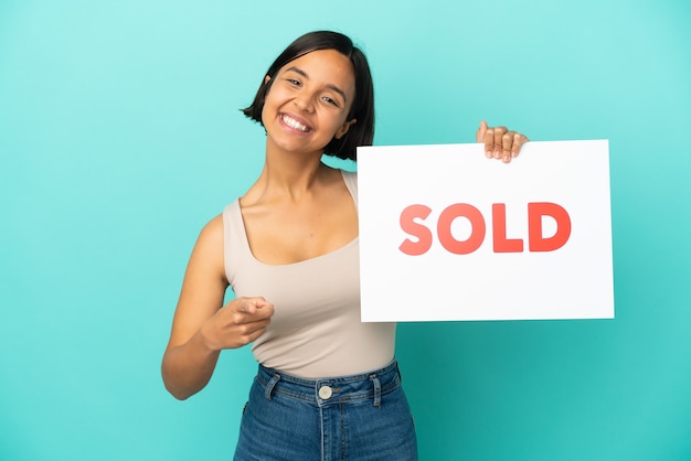 Young mixed race woman isolated on blue background holding a placard with text SOLD and pointing to the front