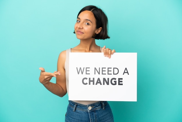 Young mixed race woman isolated on blue background holding a placard with text We Need a Change and  pointing it