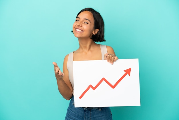 Young mixed race woman isolated on blue background holding a sign with a growing statistics arrow symbol making a deal