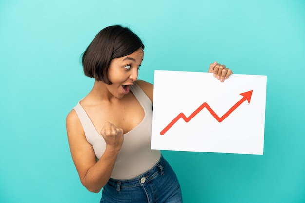 Young mixed race woman isolated holding a sign with a growing statistics arrow symbol and celebrating a victory