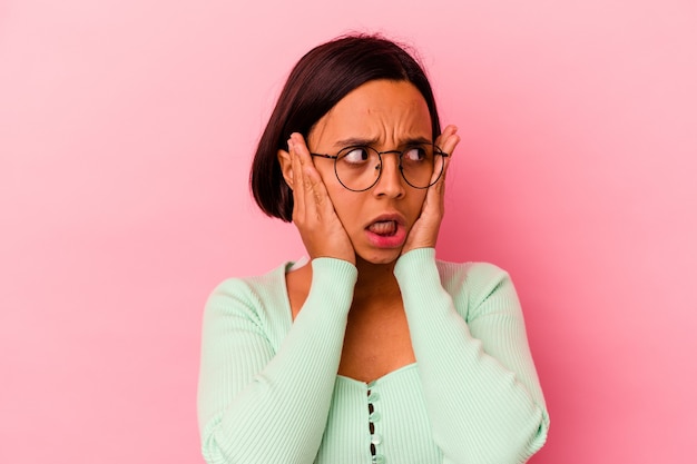 Young mixed race woman isolated on pink background covering ears with hands trying not to hear too loud sound.