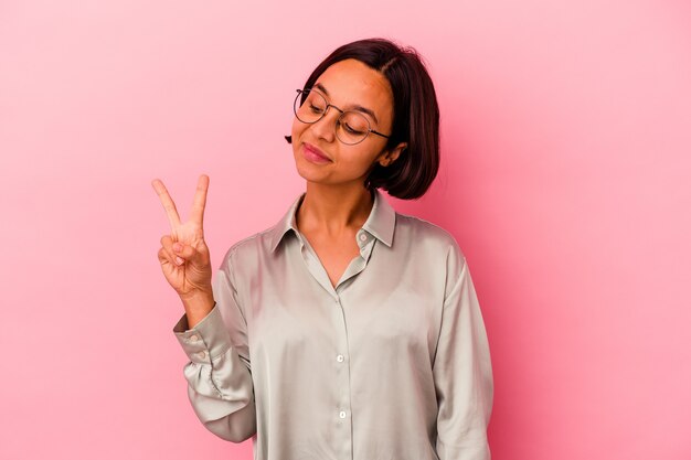 ピンクの背景に分離された若い混血の女性は、指で平和のシンボルを示して楽しくてのんきです。