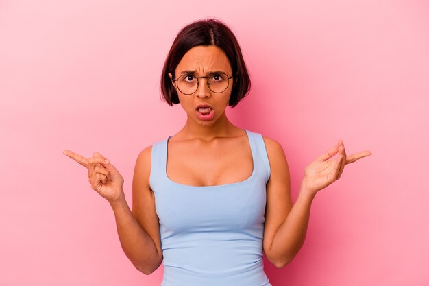 Young mixed race woman isolated on pink background pointing to different copy spaces, choosing one of them, showing with finger.