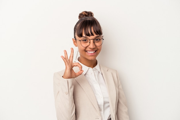 Young mixed race woman isolated on white background  winks an eye and holds an okay gesture with hand.