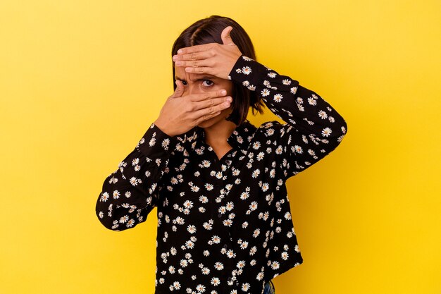 Young mixed race woman isolated on yellow background blink at the camera through fingers, embarrassed covering face.