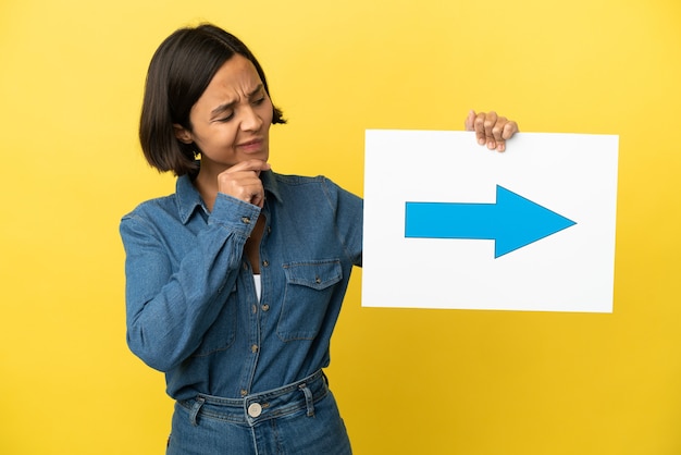 Young mixed race woman isolated on yellow background holding a placard with arrow symbol and thinking