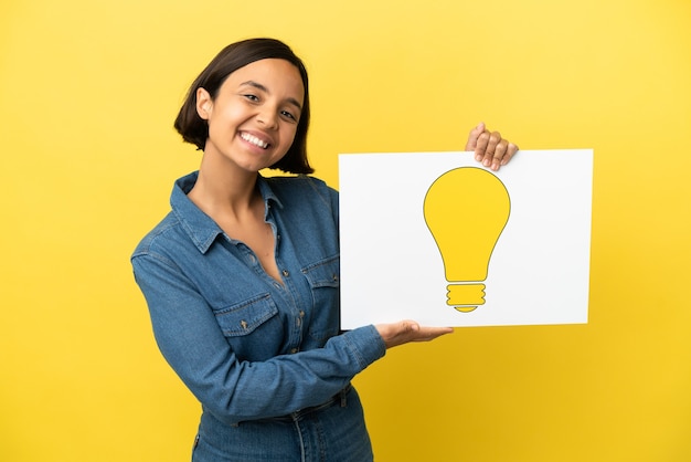 Young mixed race woman isolated on yellow background holding a placard with bulb icon with happy expression