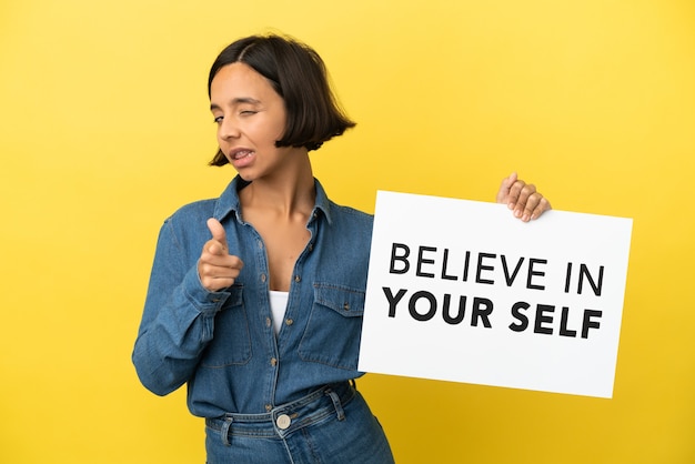 黄色の背景に分離された若い混血の女性は、テキストBelieve In Your Selfのプラカードを保持し、正面を指しています