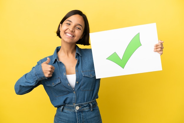 Young mixed race woman isolated on yellow background holding a placard with text Green check mark icon with thumb up