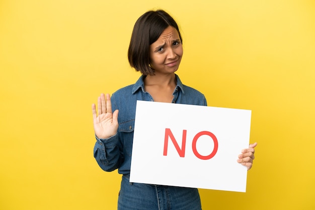 Young mixed race woman isolated on yellow background holding a placard with text no and doing stop sign