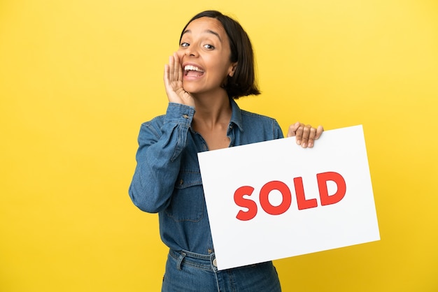 Young mixed race woman isolated on yellow background holding a placard with text SOLD and shouting