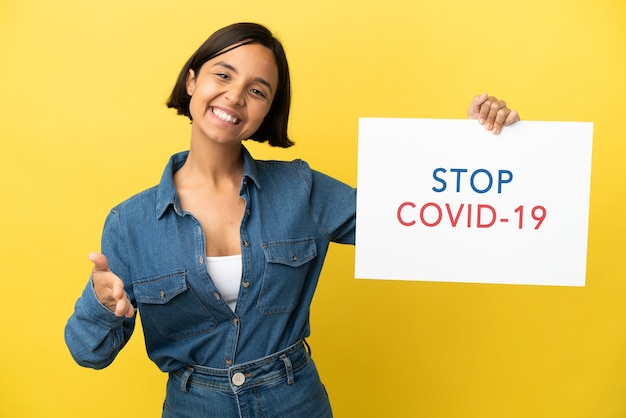 Young mixed race woman isolated on yellow background holding a placard with text Stop Covid 19 making a deal