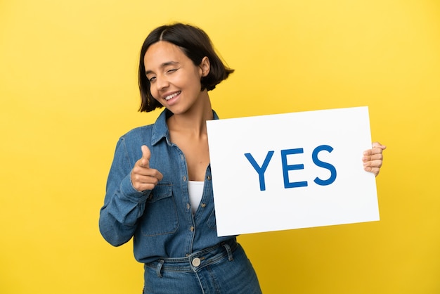 Young mixed race woman isolated on yellow background holding a placard with text YES and pointing to the front