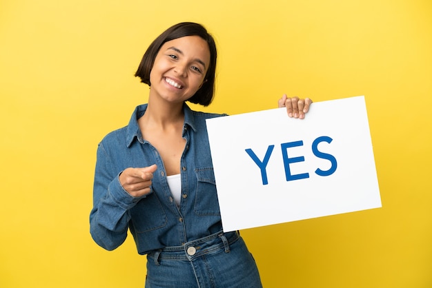 Young mixed race woman isolated on yellow background holding a placard with text YES and pointing it